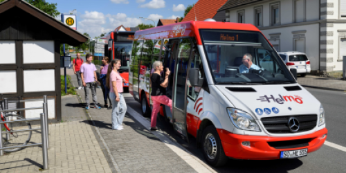 Rot-weißer Kleinbus Helmo steht an der Haltestelle. Drei Personen steigen ein.